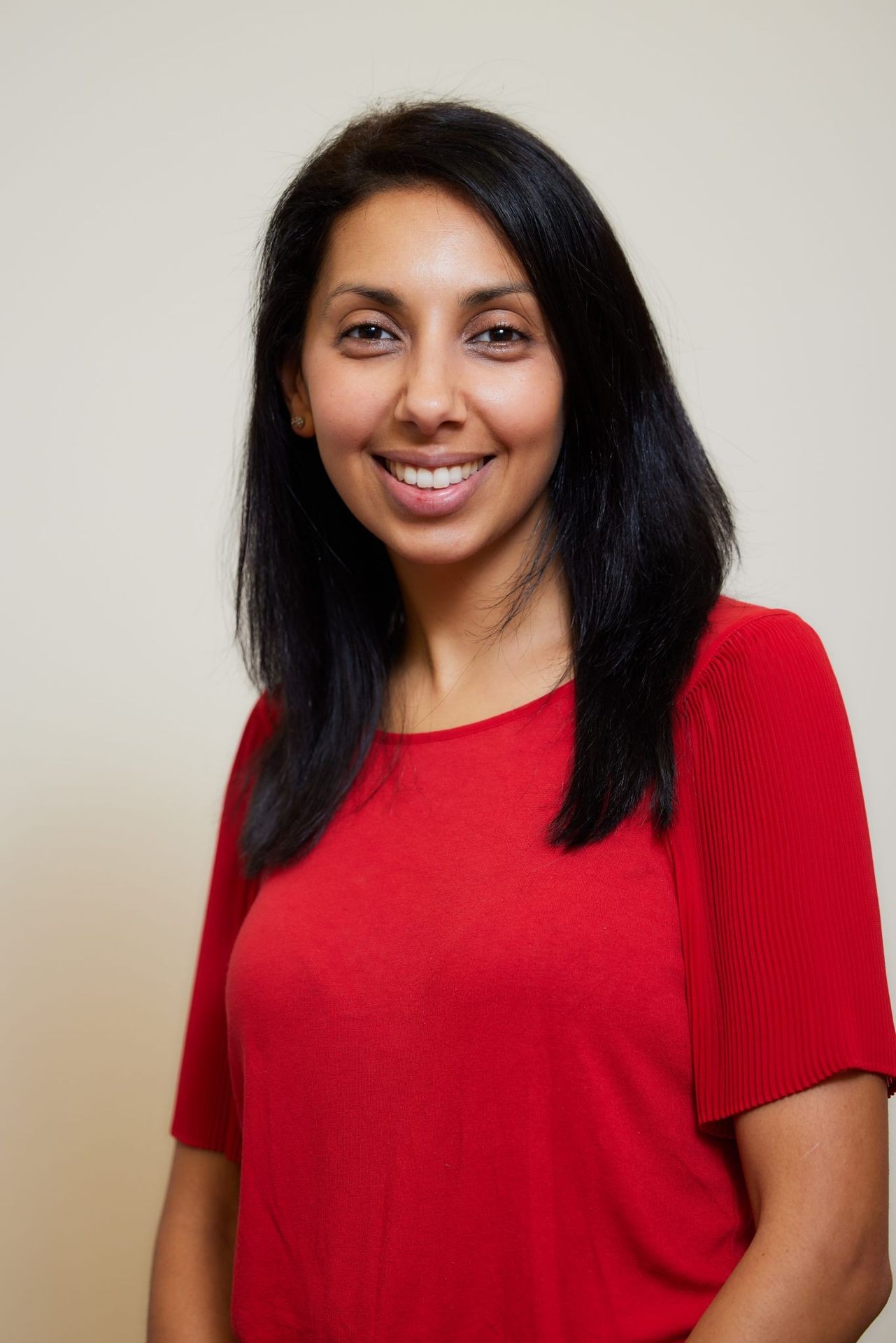 Woman with shoulder-length black hair wearing a red blouse standing against a plain background.
